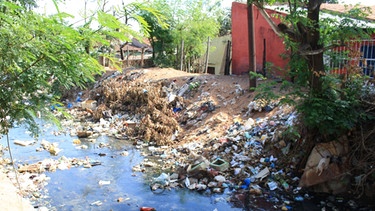Ein kleiner Fluss neben eine Holzbaracke, der voller Müll ist, auch das Ufer ist übersät mit Abfall | Bild: picture-alliance/dpa