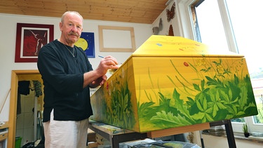 Alfred Josef Opiolka steht in seiner Werkstatt und bemalt einen Sarg.  | Bild: picture alliance / Karl-Josef Hildenbrand