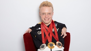 Josia Topf hat eine Gold-, eine Silber- und eine Bronzemedaille um den Hals hängen und lacht in die Kamera.  | Bild: Marie Theres Graf / Fotografschaft Erlangen 