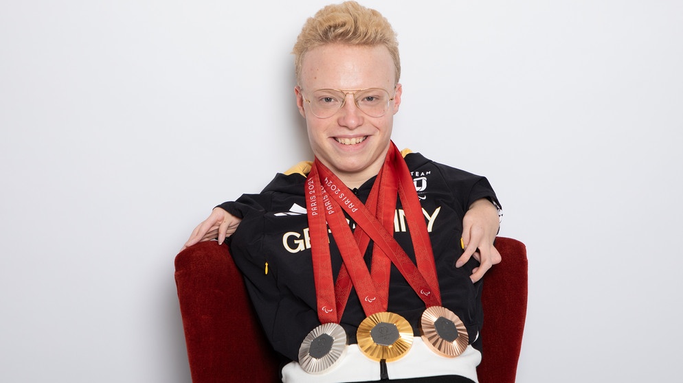 Josia Topf hat eine Gold-, eine Silber- und eine Bronzemedaille um den Hals hängen und lacht in die Kamera.  | Bild: Marie Theres Graf / Fotografschaft Erlangen 