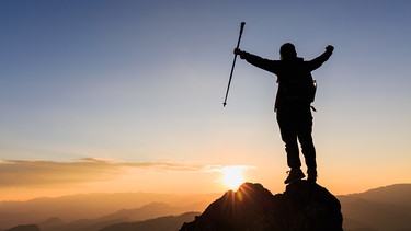 Ein Mann steht mit freudig erhobenen Armen auf einem Berggipfel, den er erklommen hat. | Bild: colourbox.com