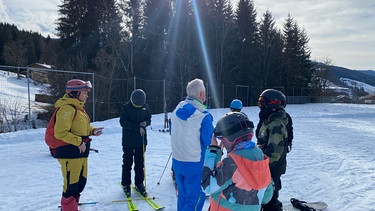 Vier Kinder auf Skiern und Snowboard stehen mit ihrer Lehrerin und ihrem Lehrer auf einer verschneiten Fläche. | Bild: BR/Joanna Thurow