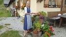 Eine Fränkin auf einer Pongauer Alm. Regina Leybold vor der Hütte auf der Klingelbergalm. Hinter ihr die Bergalm. Regina Leybold steht in einem Dirndl neben verschiedenen Blumentöpfen | Bild: BR/ Ulrike Nicola