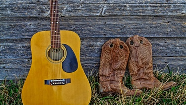 Eine Gitarre und ein Paar Stiefel lehnen an einer Holzwand | Bild: picture alliance / All Canada Photos | Murray Hayward