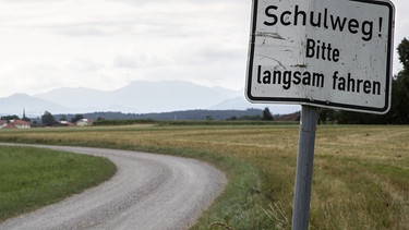 Feldweg in Landschaft, Hintergrund Berge, bewölkt, mit Schild Schulweg! Bitte langsam fahren | Bild: BR/Herbert Ebner