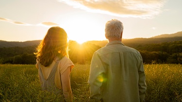 Eine Tochter steht mit ihrem Vater auf einer Wiese, während sie den Sonnenuntergang betrachten. | Bild: picture alliance / Westend61 | VITTA GALLERY