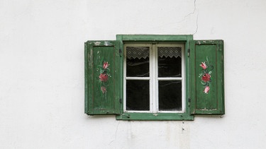 Ein altes Fenster mit grünen Fensterläden, die mit Blumen bemalt sind. | Bild: stock.adobe.com/hanneliese