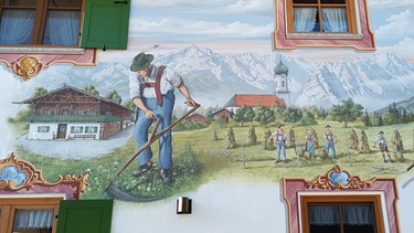 Alpine Fassadenkunst. Man sieht einen Teil eines Hauses im Alpenvorland das bemalt ist mit Bergen, Himmel, Bauern beim Heumachen, die Fenster sind umrahmt | Bild: Bettina Weiz