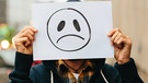 Eine Person hält ein Blatt Papier mit einem traurigen Smilie vor das Gesicht | Bild: BR / Lisa Hinder