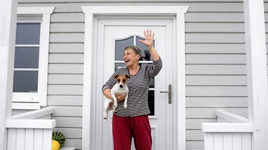 Eine Frau steht mit ihrem Hund in der Hand vor ihrer Haustür und winkt jemandem zu | Bild: picture alliance / Westend61 | Katharina Mikhrin