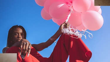 Die Rapperin Kokonelle ist halb sitzend, halb liegend zu sehen. Sie trägt ein rotes Outfit und hält rosafarbende Ballons in der Hand.  | Bild: Andreas Roppel