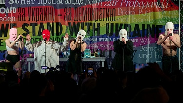 Beim Konzert der Band Pussy Riot gegen den Krieg in der Ukraine am Brandenburger Tor stehen Bandmitglieder mit Stoffmasken auf der Bühne. Auf der Fahne hinter ihnen steht unter anderem "We stand with Ukraine", also "wir stehen mit der Ukraine". | Bild: picture alliance/dpa | Michael Ukas