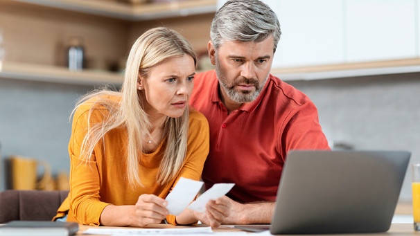 Ein Paar erledigt die private Buchhaltung in der Küche, vor sich ein Laptop. Die Frau hat zwei Kassenzettel in der Hand. | Bild: mauritius images / Prostock-studio / Alamy / Alamy Stock Fotos