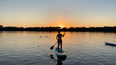 Gemütlich in den Sommerabend: Eine junge Frau auf einem Stand-Up Paddle bei Sonnenuntergang. | Bild: BR/Annette Goossens