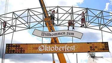 Arbeiter klettern auf dem Gerüst des Eingangstors des Oktoberfests vor weißblauem Himmel während des Wiesn-Aufbau auf der Theresienwiese.  | Bild: dpa-Bildfunk/Felix Hörhager