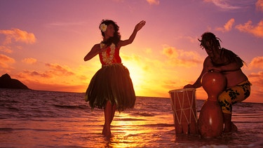 Hula dancer performing with drummer on beach at sunrise in background Hawaii. | Bild: picture alliance / Design Pics / Pacific Stock | Tomas del Amo