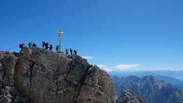 Zugspitze mit Bergsteigern und Gipfelkreuz. 2.962 Meter ragt Deutschlands höchster Berg in die Höhe: die Zugspitze. Vor genau 200 Jahren hat sie ein Tiroler zum ersten Mal bestiegen - zumindest offiziell.  | Bild: Manuel Rauch BR