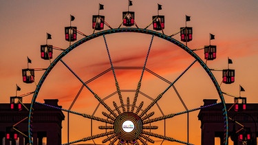 10.08.2021, Bayern, München: Ein Riesenrad leuchtet am späten Abend auf dem Königsplatz vor der Kulisse der Propyläen, das dort für den "Sommer in der Stadt" aufgebaut ist. Mit dem Programm sollen in ganz München Fahrgeschäfte und andere Attraktionen aufgebaut werden, um die Schausteller zu unterstützen. Wegen der abermaligen Absage des Oktoberfestes haben viele Betriebe finanzielle Einbußen. (Langzeitbelichtung) Foto: Peter Kneffel/dpa +++ dpa-Bildfunk +++ | Bild: dpa-Bildfunk/Peter Kneffel