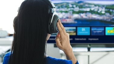 Eine Frau mit Kopfhörern blickt auf ein modernes Smart-TV. Sie hört die Tonspur Audiodeskription zur laufenden Sendeung. | Bild: BR/Lisa Hinder; Collage/Petra Decker