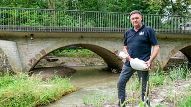 Michael Lukacz steht mit einem Sandsack neben einem Fluss, der Schwarzach, und einer Brücke und blickt besorgt in die Kamera. | Bild: BR/Florian Barnikel
